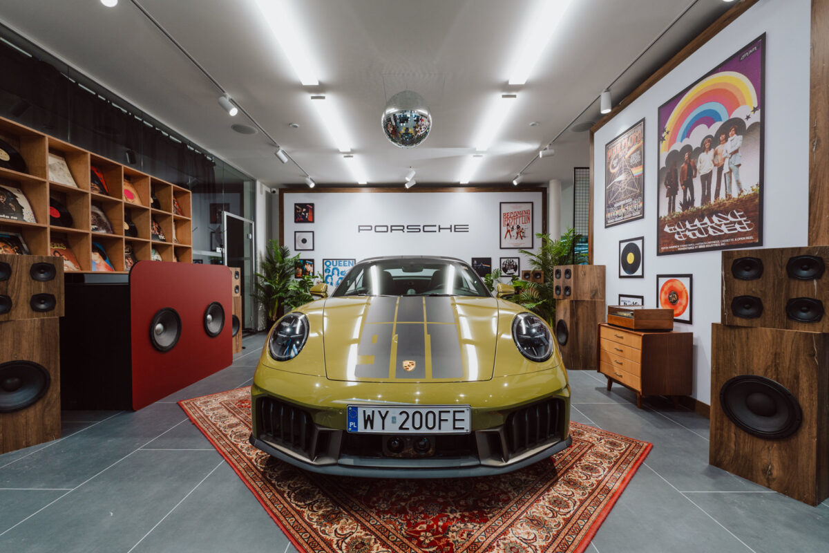 Ein grüner Porsche-Sportwagen parkt im Inneren auf einem gemusterten Teppich, umgeben von Vintage-Lautsprechern, Schallplatten und musikbezogenem Dekor - perfekt für eine Showroom-Eröffnung, mit Schallplattenhüllen und einer von der Decke hängenden Discokugel.