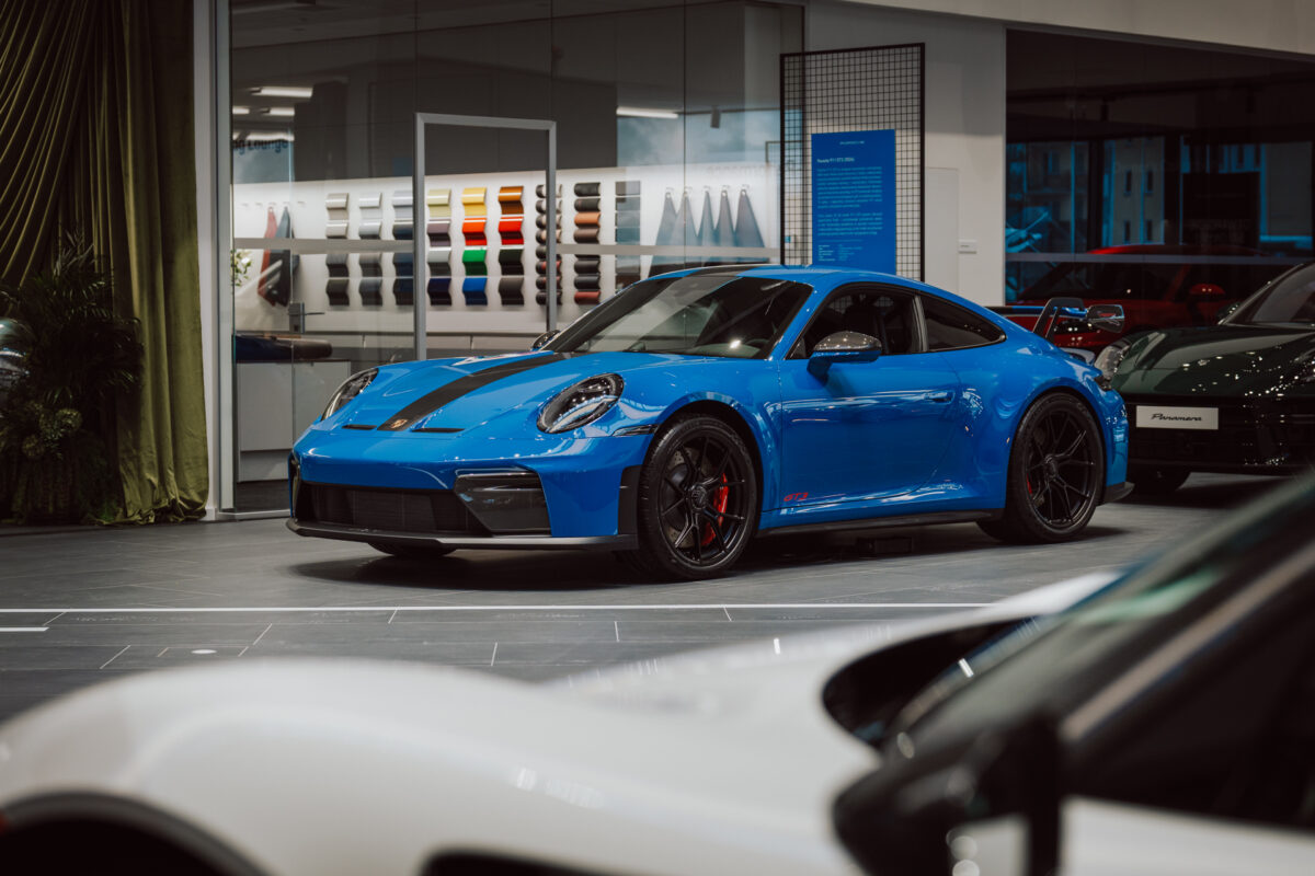 Ein blauer Porsche Sportwagen wird während der Eröffnung des Showrooms im modernen Showroom ausgestellt, mit Farbmustern an der Wand. Andere Fahrzeuge und Lounge-Dekor sind teilweise im Vorder- und Hintergrund zu sehen. 