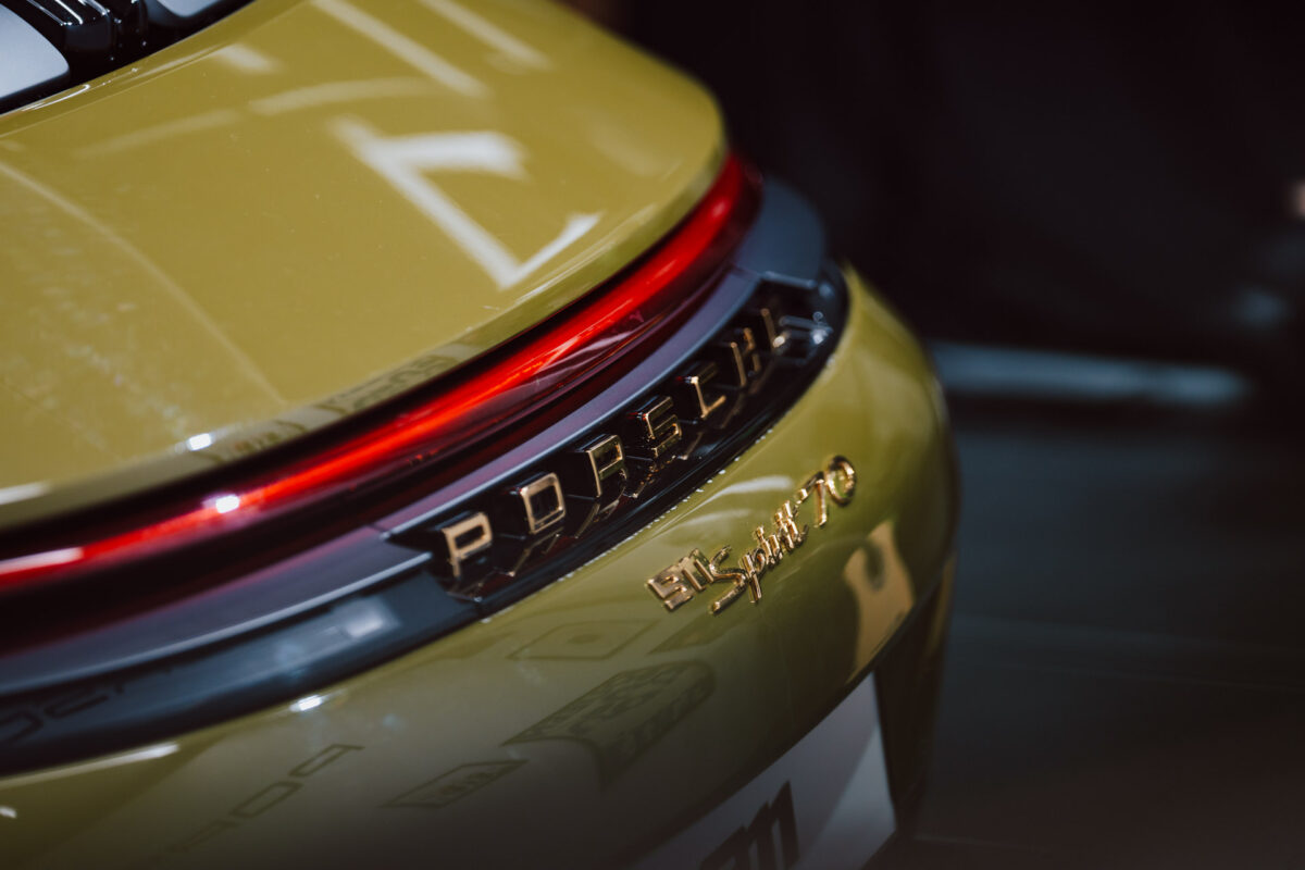 Nahaufnahme des Hecks eines goldenen Porsche-Sportwagens in einem offenen Ausstellungsraum. Zu sehen sind das Porsche-Logo und die '911 S/T 70'-Plakette, elegante Rückleuchten und Spiegelungen auf der glänzenden Oberfläche.