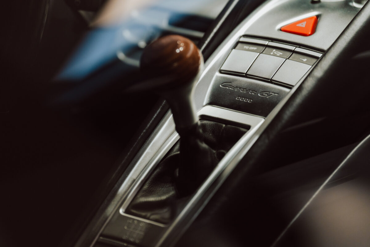 Nahaufnahme des Schalthebels und der Mittelkonsole des Fahrzeugs mit den Tasten und dem roten dreieckigen Notrufknopf. Auf der Konsole ist ein Modell des 'Carrera GT' eingraviert - ideal für die Präsentation bei einer Ausstellungseröffnung. 