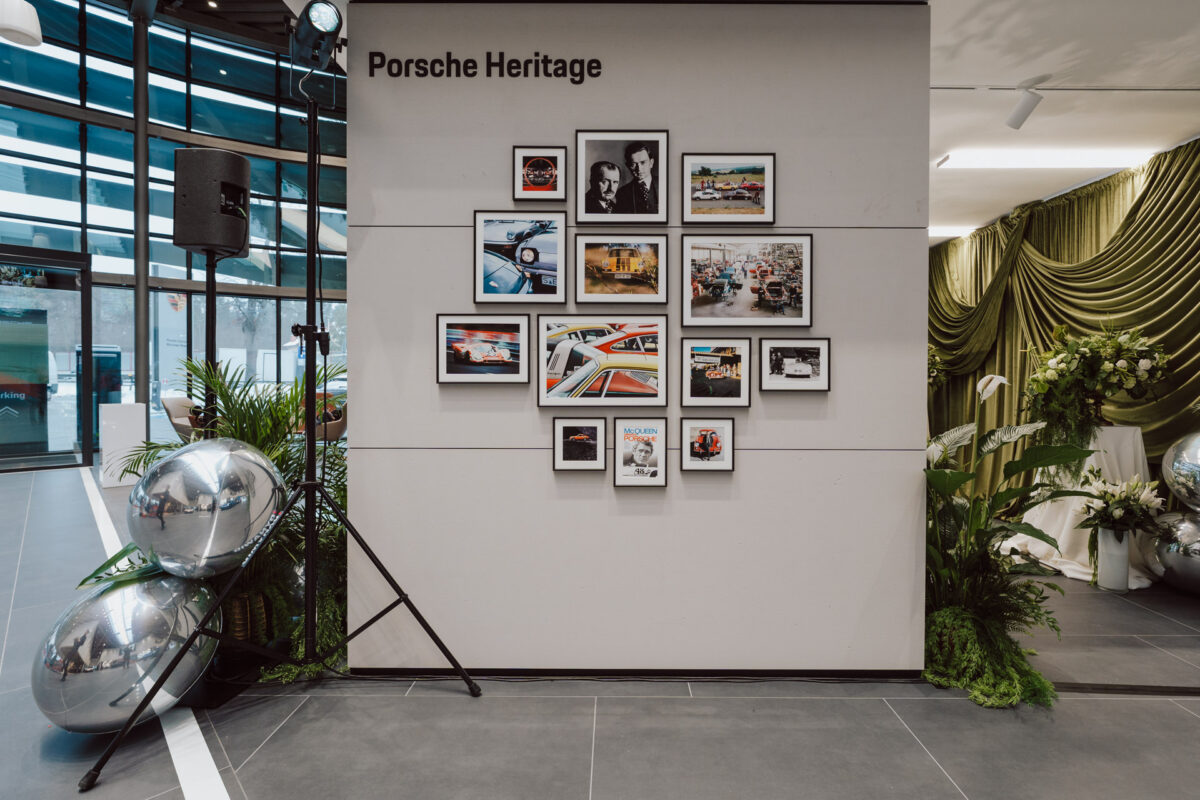 Die Wandausstellung mit dem Titel 'Porsche Heritage' zeigt eine Collage aus gerahmten Fotografien und Kunstwerken, hauptsächlich Porsche-Fahrzeuge, die die Eröffnung des Showrooms in einem modernen Wohnzimmer mit Pflanzen, metallischen Dekokugeln und einem grün drapierten Hintergrund feiern.