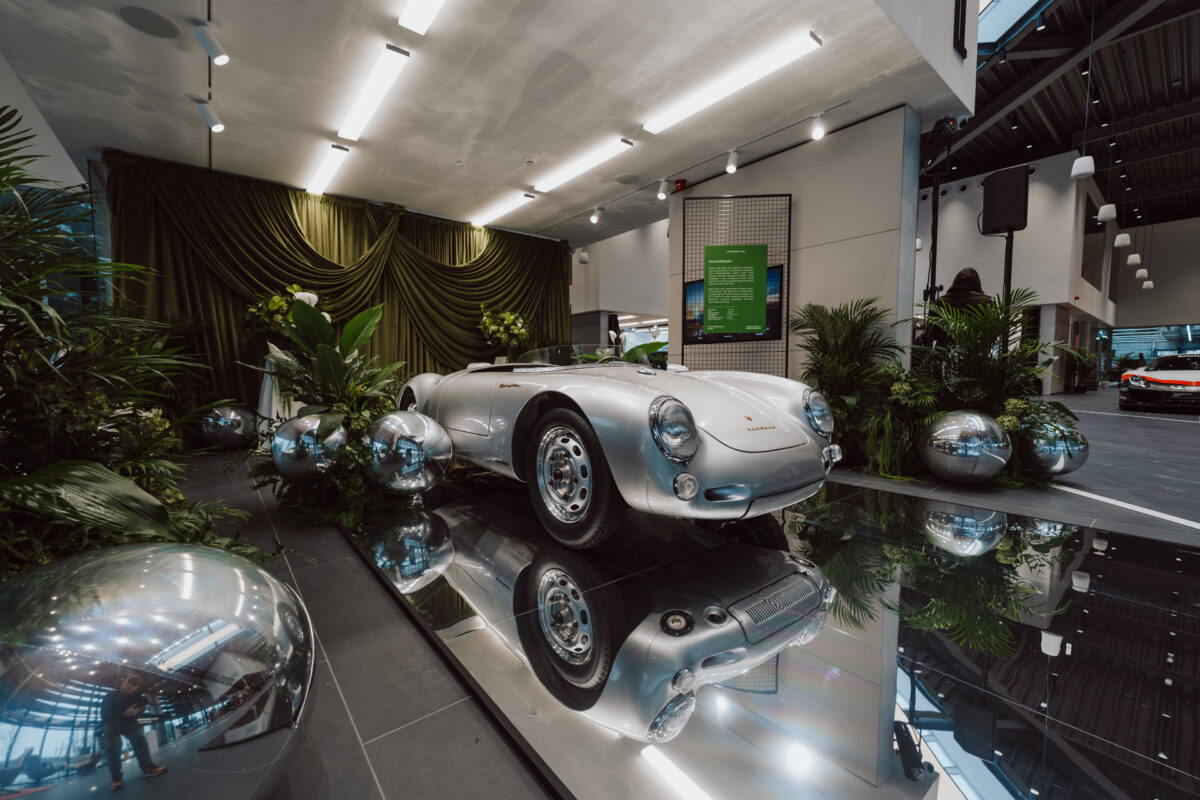 Der klassische silberne Sportwagen steht in dem Raum auf einem spiegelnden Boden, während sich der Ausstellungsraum öffnet, umgeben von grünen Pflanzen, glänzenden Metallkugeln, grünen Vorhängen und moderner Deckenbeleuchtung.