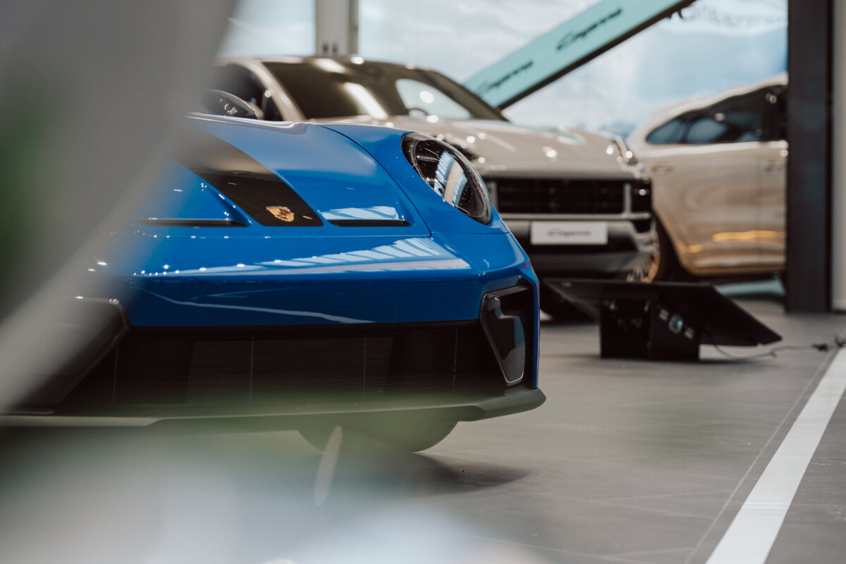 Nahaufnahme der vorderen linken Seite eines blauen Sportwagens im Showroom während der Eröffnung des Showrooms, mit zwei weiteren Luxusautos, einem beigen und einem goldenen, unscharf vor einem hellen natürlichen Hintergrund.