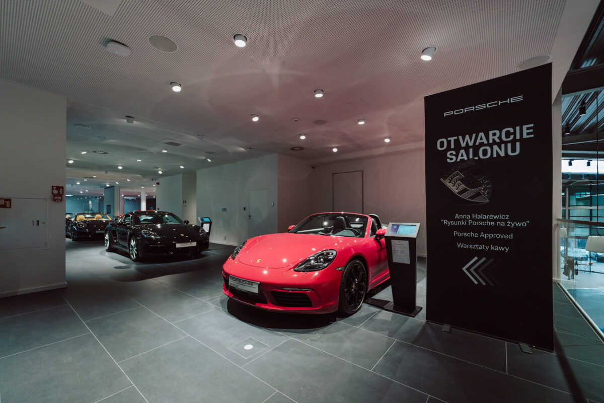Ein moderner Autohaus mit einem roten Porsche Sportwagen im Vordergrund, einem schwarzen Porsche im Hintergrund und der Bildunterschrift 'Porsche Showroom Opening', die die Details der Eröffnungsveranstaltung auf Polnisch hervorhebt.