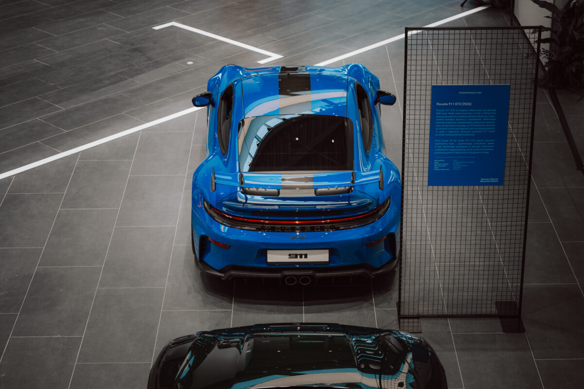 Ein hellblauer Sportwagen mit schwarzen Rennstreifen und einem großen Heckspoiler steht in der Öffnung des Ausstellungsraums, von oben gesehen neben dem Informationsschild. Der grau geflieste Boden unterstreicht die Eleganz der Szene. 