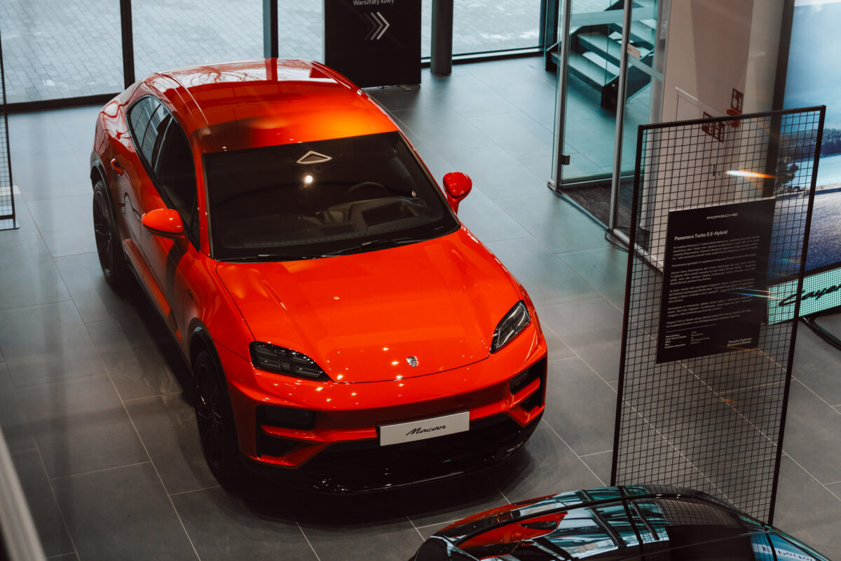 Der leuchtend rote Porsche Macan ist in einem modernen Ausstellungsraum mit großen Glasfenstern und einem eleganten Fliesenboden ausgestellt, bereit für die Eröffnung des Showrooms. In der Nähe befinden sich Informationsstände. 