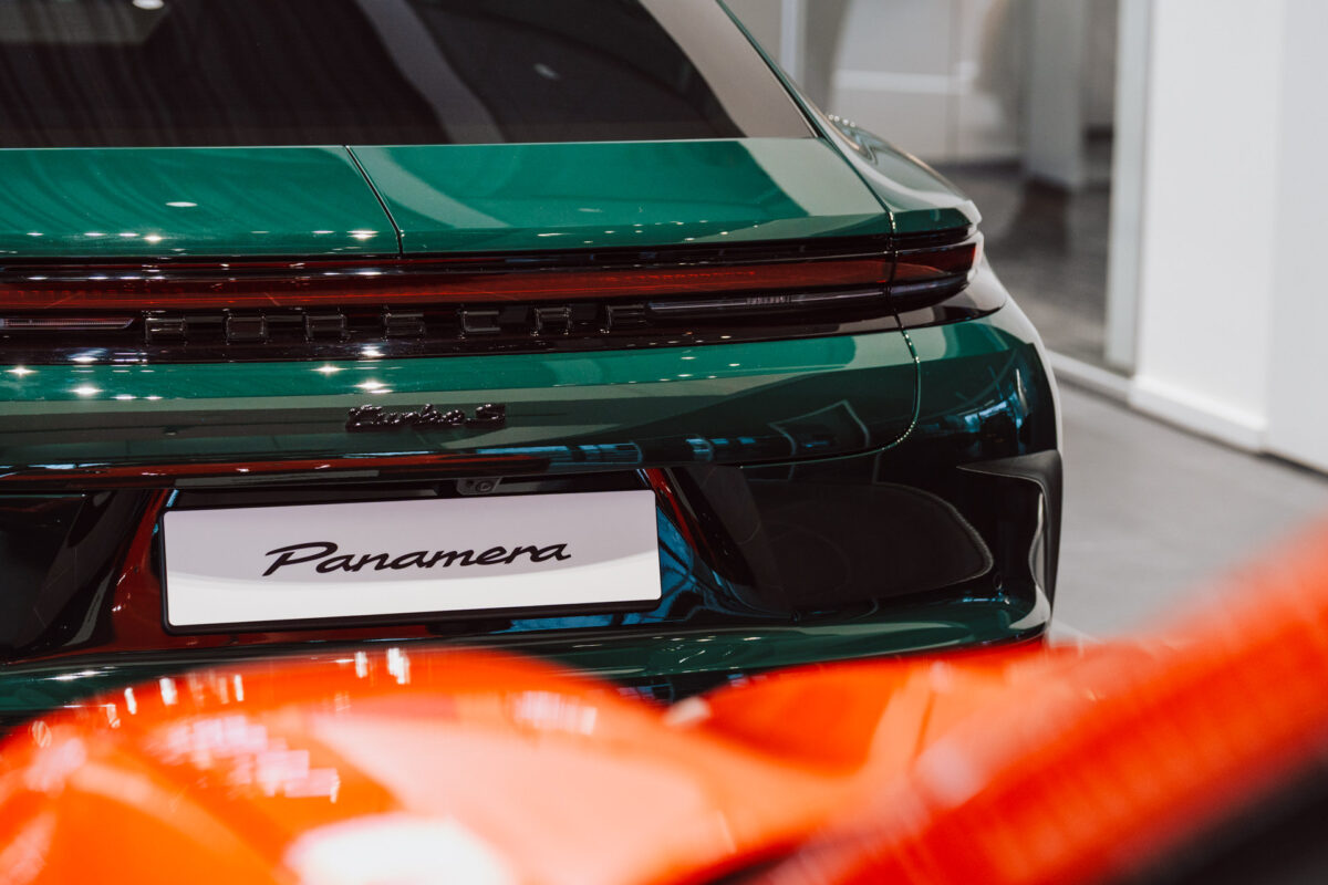 Nahaufnahme des Hecks eines dunkelgrünen Porsche Panamera, wobei die Panamera-Plakette und das Rücklicht hervorgehoben werden, mit dem unscharfen roten Auto im Vordergrund während der Eröffnung des Showrooms.
