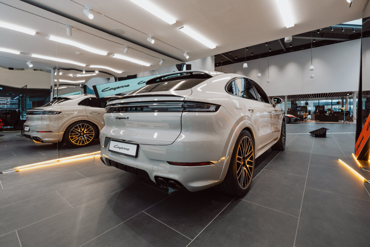 Der weiße Porsche Cayenne wird im Inneren auf einem grau gefliesten Boden mit heller Beleuchtung präsentiert, der bei der Eröffnung des Showrooms vorgestellt wurde. Der Wagen ist von hinten zu sehen, wobei sein schlankes Design und die goldenen Leichtmetallfelgen zur Geltung kommen, während man sein Spiegelbild in einem Wandspiegel sieht. 