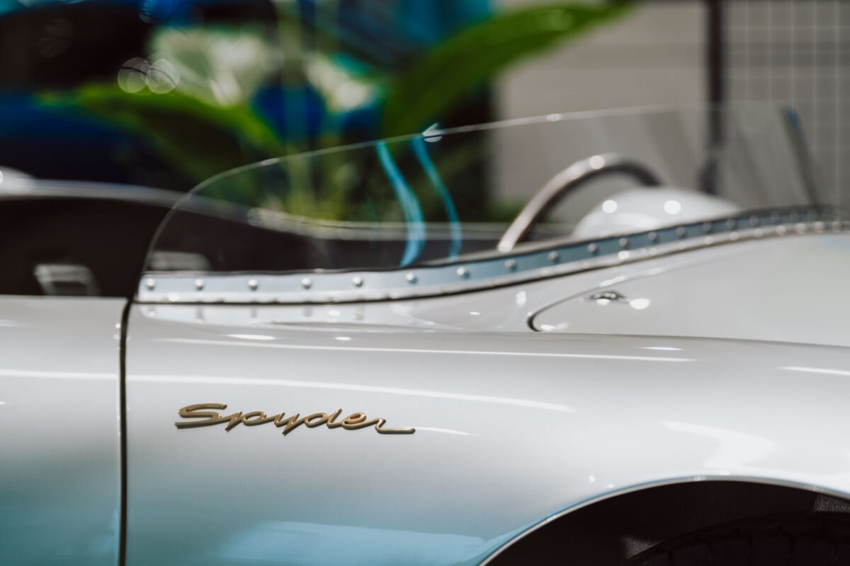 Nahaufnahme eines klassischen silbernen Autos mit goldenem 'Spyder'-Schriftzug an der Seite, das in einem offenen Ausstellungsraum präsentiert wird. Man sieht die kleine Windschutzscheibe und die genieteten Zierleisten, mit unscharfen Grün- und Blautönen im Hintergrund.
