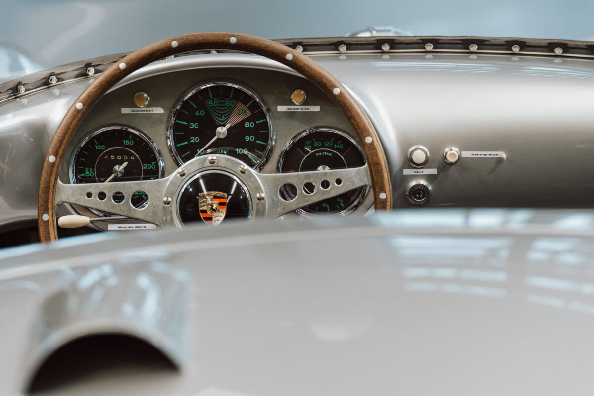 Nahaufnahme des klassischen Porsche Armaturenbretts mit analogen Anzeigen, Holzlenkrad mit Porsche Wappen und Metalldetails - aufgenommen bei der Eröffnung des Showrooms, vom Fahrersitz aus gesehen.