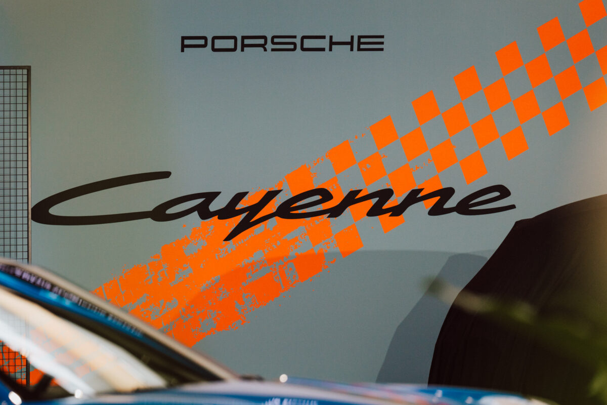 Eine Wand mit großem 'Porsche Cayenne'-Schriftzug und orangefarbenen karierten Grafiken zur Feier der Eröffnung des Showrooms. Die Vorderseite des blauen Autos erscheint unten im Bild. 