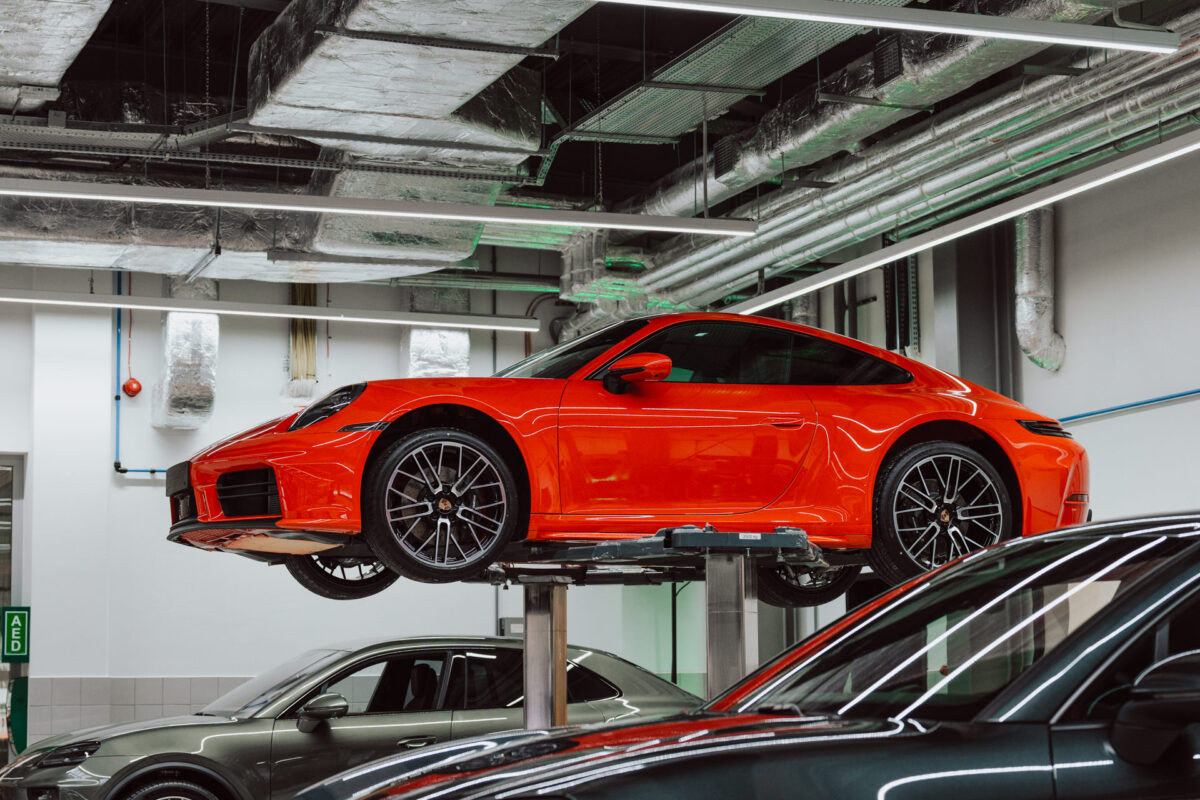 Ein roter Sportwagen wird in einer modernen Autowerkstatt während der Eröffnung des Showrooms auf einem hydraulischen Wagenheber angehoben, wobei die Deckenrohre über dem Boden sichtbar sind. Andere Fahrzeuge sind in der Nähe in der gut beleuchteten Garage geparkt. 