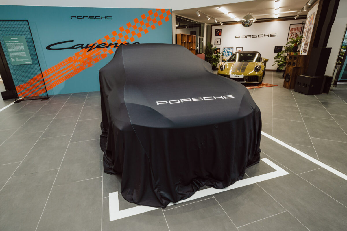 Ein abgedeckter Porsche, der in einem offenen Ausstellungsraum ausgestellt wird, mit einem Porsche Taycan und bunten Wandgrafiken im Hintergrund. Die schwarze Abdeckung ist mit dem Porsche-Logo versehen. 