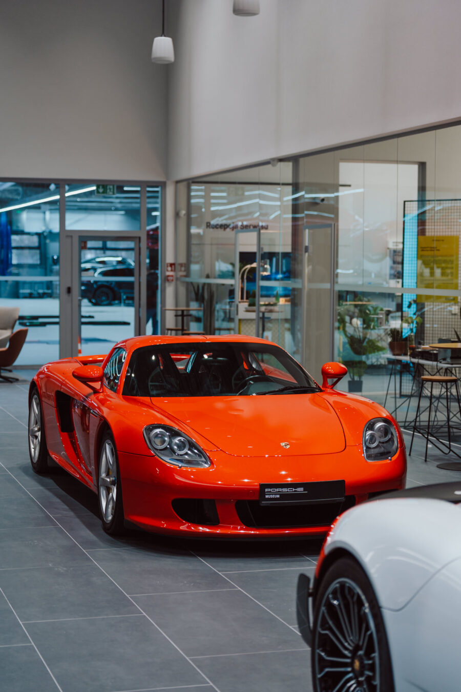 Der leuchtend rote Porsche Carrera GT Sportwagen wird während der Eröffnung des Showrooms in einem modernen Showroom mit Glaswänden und elegantem Interieur ausgestellt; im Vordergrund ist ein Teil des weißen Autos zu sehen.
