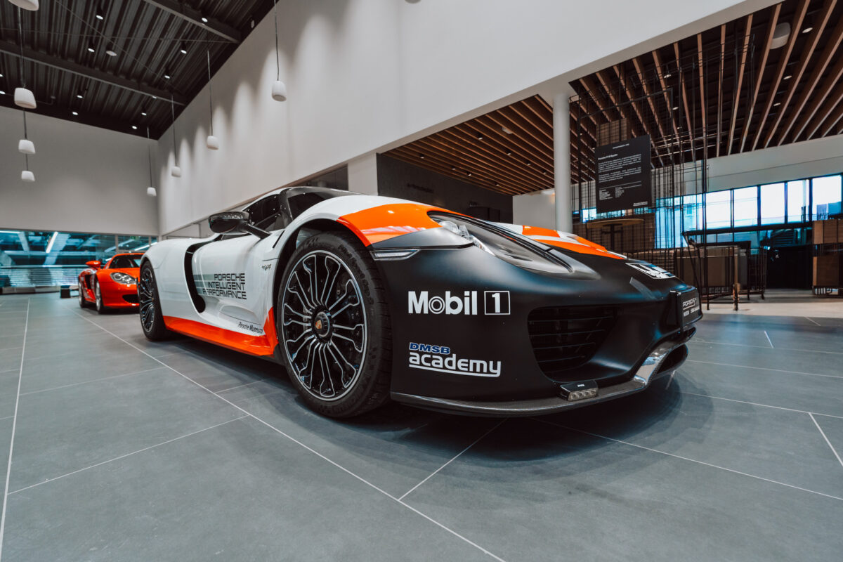 Ein schwarz-weiß-roter Porsche-Sportwagen mit Rennaufklebern ist während der Eröffnung des Showrooms in dem Raum auf der Etage ausgestellt, während ein weiteres rotes Auto im Hintergrund unter modernen Hängelampen zu sehen ist.