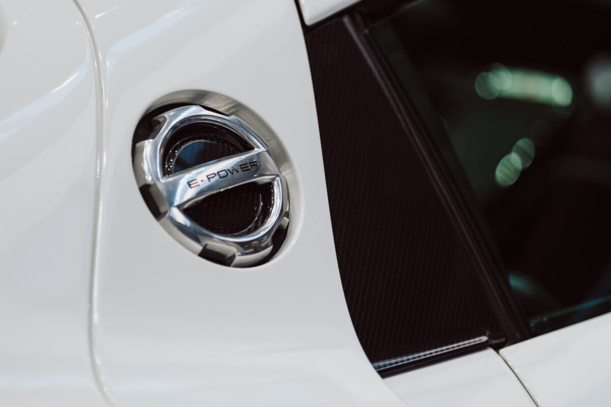 Eine Nahaufnahme des silbernen Ladeanschlusses mit dem Schriftzug 'E-POWER' auf einem weißen Fahrzeug, der das schlanke, moderne Design und die glänzende Oberfläche hervorhebt - perfekt für die Eröffnung eines Ausstellungsraums, in dem innovative Automobiltechnologie präsentiert wird.