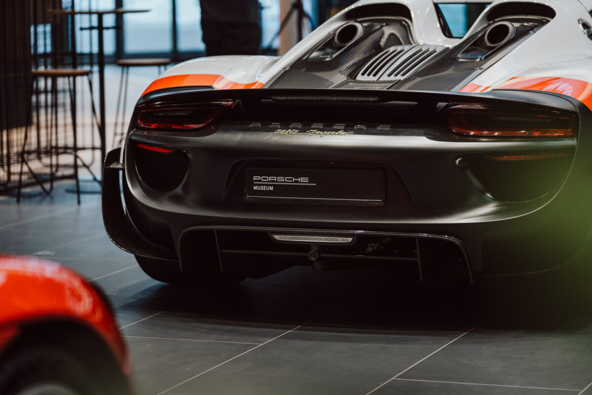Rückansicht des weiß-orangefarbenen Porsche 918 Spyder, der im Porsche Museum ausgestellt ist. Das schnittige Design, die markanten Rückleuchten und die auffälligen Motorentlüftungen werden bei der Eröffnung des Showrooms gezeigt.