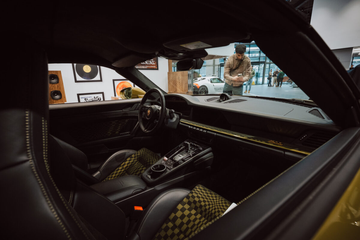 Blick auf das schwarze Lederinterieur eines Autos mit gelben Nähten und karierten Sitzen, aufgenommen von der Beifahrerseite während der Eröffnung des Ausstellungsraums; eine Person steht in der Nähe im Ausstellungsraum mit Kunstwerken und einem anderen Auto im Hintergrund.