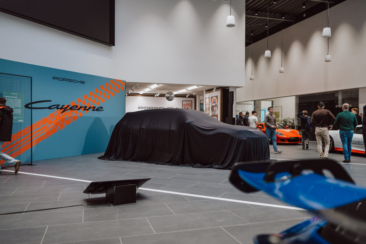 Ein überdachtes Auto, das während der Eröffnung eines Ausstellungsraums in einem Porsche-Autohaus ausgestellt wird, mit Menschen, die in der Nähe stehen. Das blau-orangefarbene 'Cayenne'-Display ist im Hintergrund zu sehen, und ein weiteres orangefarbenes Auto ist im Ausstellungsraum zu sehen. 