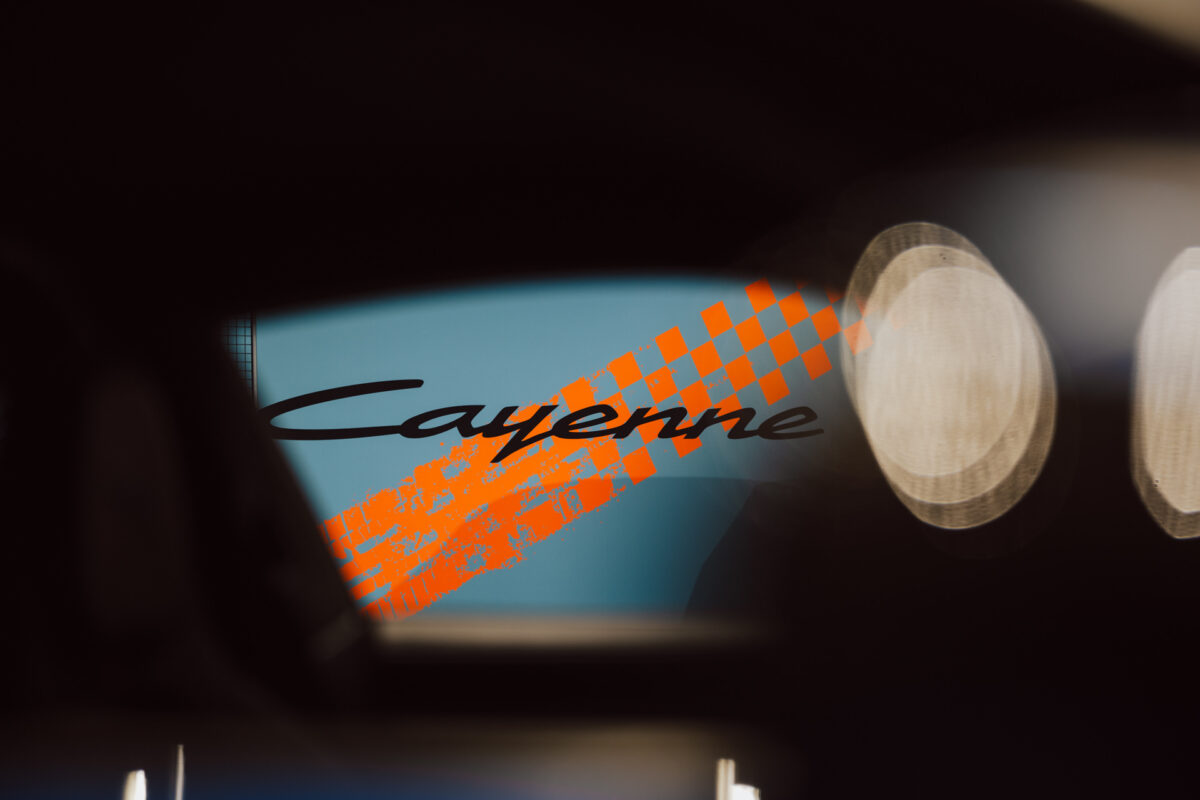 Eine Nahaufnahme des Armaturenbretts des Fahrzeugs mit dem Schriftzug 'Cayenne' und einem orangefarbenen karierten Streifen, der teilweise mit blauen und schwarzen Hintergrundelementen verschwimmt - perfekt, um die Aufregung bei der Eröffnung des Showrooms einzufangen.