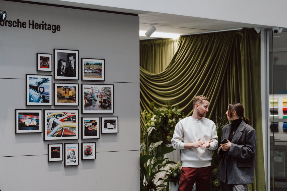 Ein Mann und eine Frau unterhalten sich bei der Eröffnung des Showrooms an einer Wand mit gerahmten Porsche Fotos und dem Text 'Porsche Heritage'. Grüne Vorhänge und Pflanzen schmücken das moderne Interieur. 
