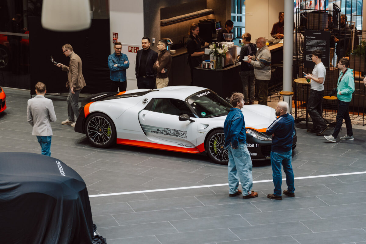 Bei der Eröffnung des Showrooms steht eine Gruppe von Menschen um einen weißen Porsche Sportwagen mit orangefarbenen Akzenten herum und unterhält sich. Einige machen Fotos, andere schauen zu oder unterhalten sich in dem modernen Ausstellungsraum. 