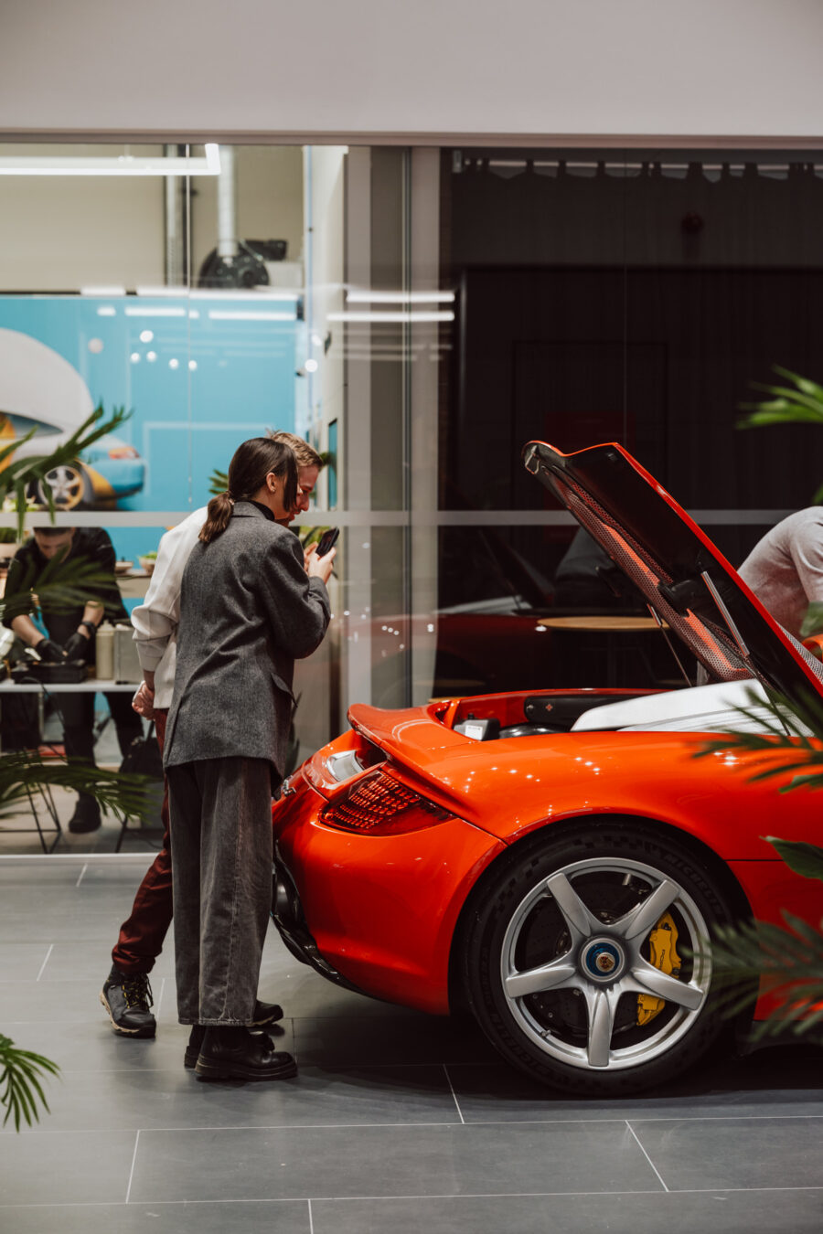 Zwei Personen untersuchen den offenen Motorraum eines roten Sportwagens, der in einem offenen Ausstellungsraum ausgestellt ist. Im Hintergrund sind eine moderne Einrichtung und ein Poster des Wagens zu sehen.
