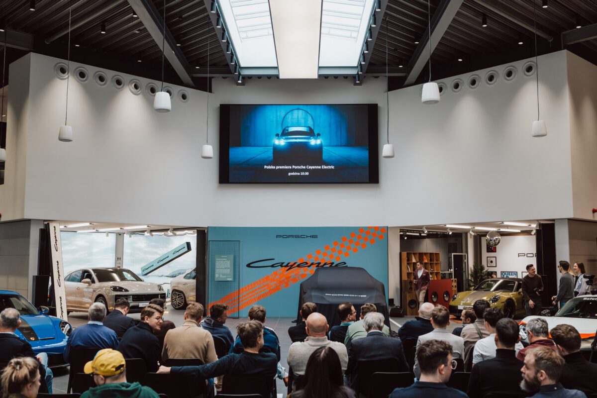 Bei der Eröffnungsveranstaltung des Showrooms sitzt eine Menschenmenge vor einem Bildschirm, auf dem ein Porsche-Fahrzeug zu sehen ist. Auf beiden Seiten werden unter einer modernen, leuchtenden Decke zwei Porsche-Fahrzeuge präsentiert.