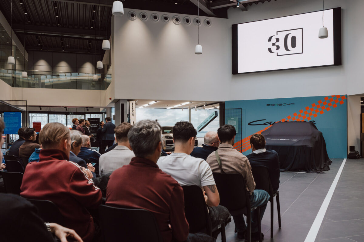 Eine Gruppe von Menschen sitzt bei einer Party in einem modernen Autohaus während der Eröffnung des Showrooms vor der Bühne. Auf einem großen Bildschirm wird '30' über einem teilweise überdachten Porsche angezeigt, eingebettet in elegante Architektur und Glaswände. 