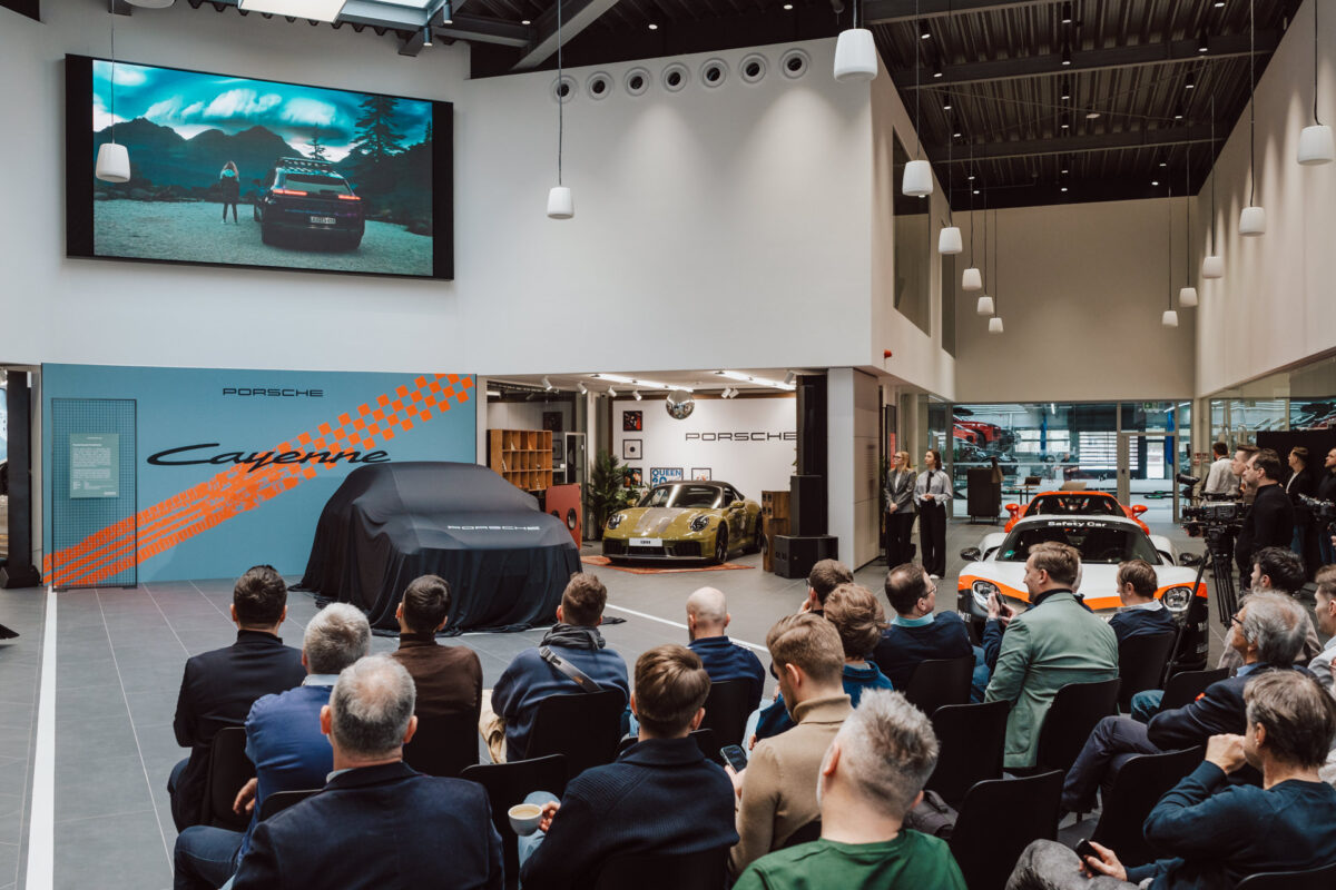 Während der Eröffnung des Showrooms sitzt eine Gruppe von Menschen vor der Bühne im modernen Showroom und schaut auf einen großen Bildschirm über dem überdachten Auto, auf dem im Hintergrund mehrere Porsche zu sehen sind.