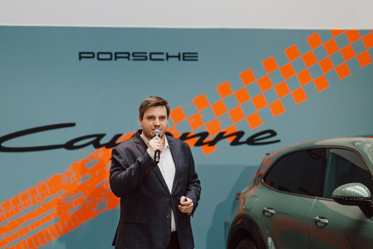 Bei der Eröffnung des Showrooms spricht ein Mann in einer Jacke in ein Mikrofon neben einem grünen Auto, vor dem Hintergrund des Porsche-Logos, eines orangefarbenen Schachbretts und der Worte 'Cayenne'.