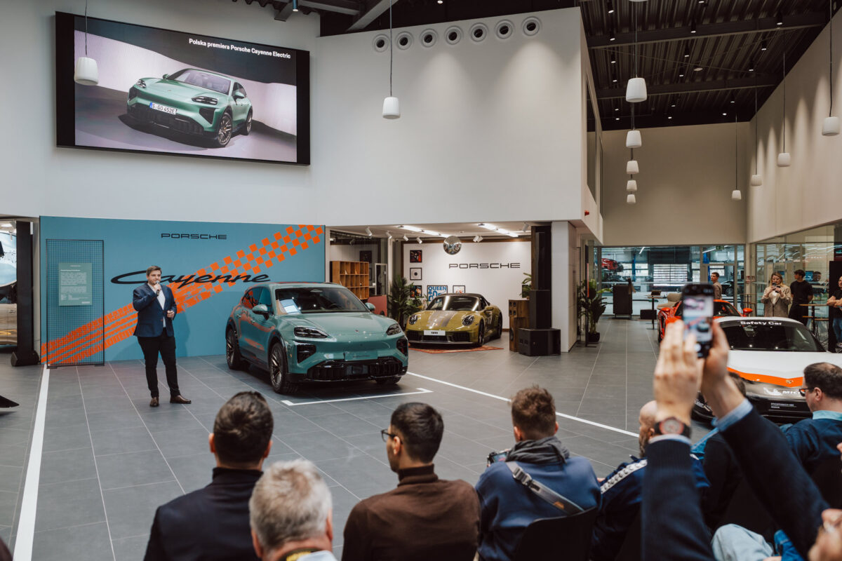 Während der Eröffnung des Showrooms steht der Moderator neben einem grünen Porsche Cayenne im Showroom und spricht zum Publikum. Die Besucher schauen zu und machen Fotos, während im Hintergrund andere Porsches und ein großer Bildschirm zu sehen sind, auf dem das Auto zu sehen ist. 