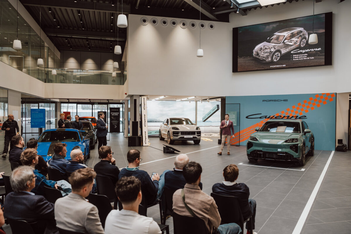 Ein Moderator spricht zu den Zuhörern in einem modernen Ausstellungsraum bei der Eröffnung des Showrooms, in dem drei Porsche Fahrzeuge ausgestellt sind. Auf dem großen Bildschirm darüber werden Bilder und Details des Porsche Cayenne gezeigt, während die Anwesenden aufmerksam zuhören. 