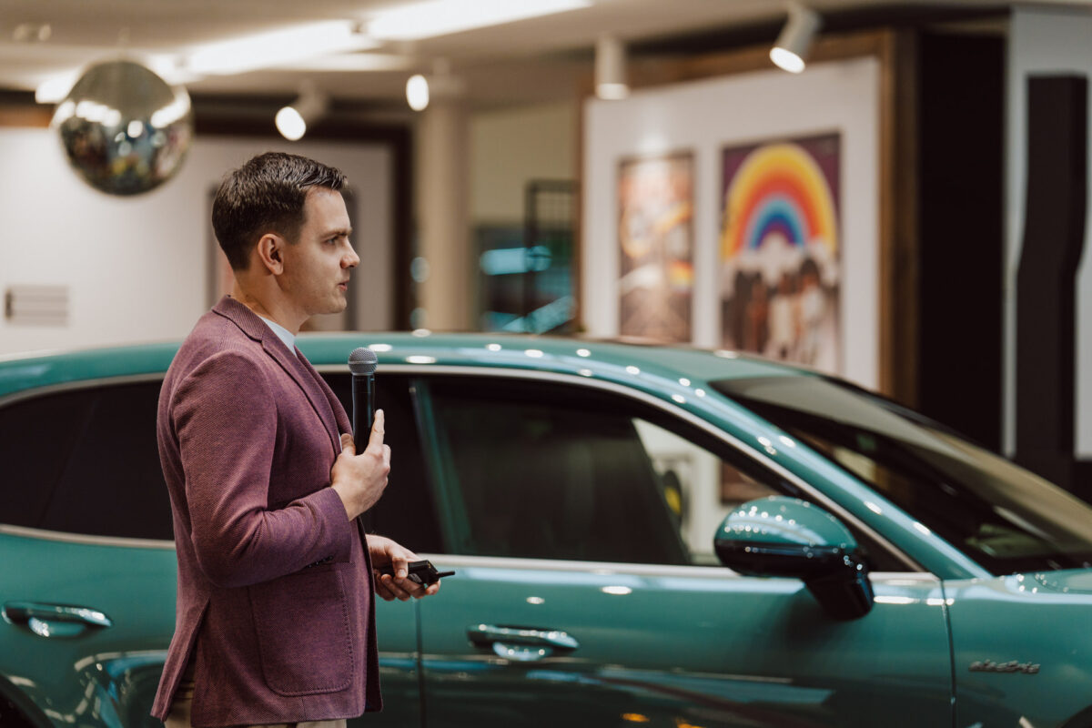 Bei der Eröffnung des Showrooms hält ein Mann in einer lila Jacke ein Mikrofon in der Hand und spricht vor einem türkisfarbenen Auto im Innenraum, dessen bunte Grafiken und Scheinwerfer den Hintergrund beleuchten.