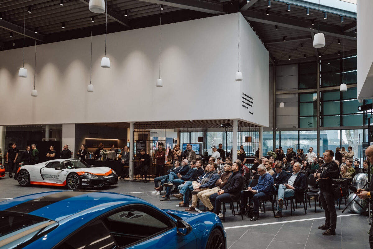Bei der Eröffnung sitzt eine Gruppe von Menschen vor dem modernen Ausstellungsraum, in dem Sportwagen ausgestellt sind. Große Fenster, hohe Decken und Hängelampen sind zu sehen, während einige im Hintergrund stehen und das Geschehen beobachten. 