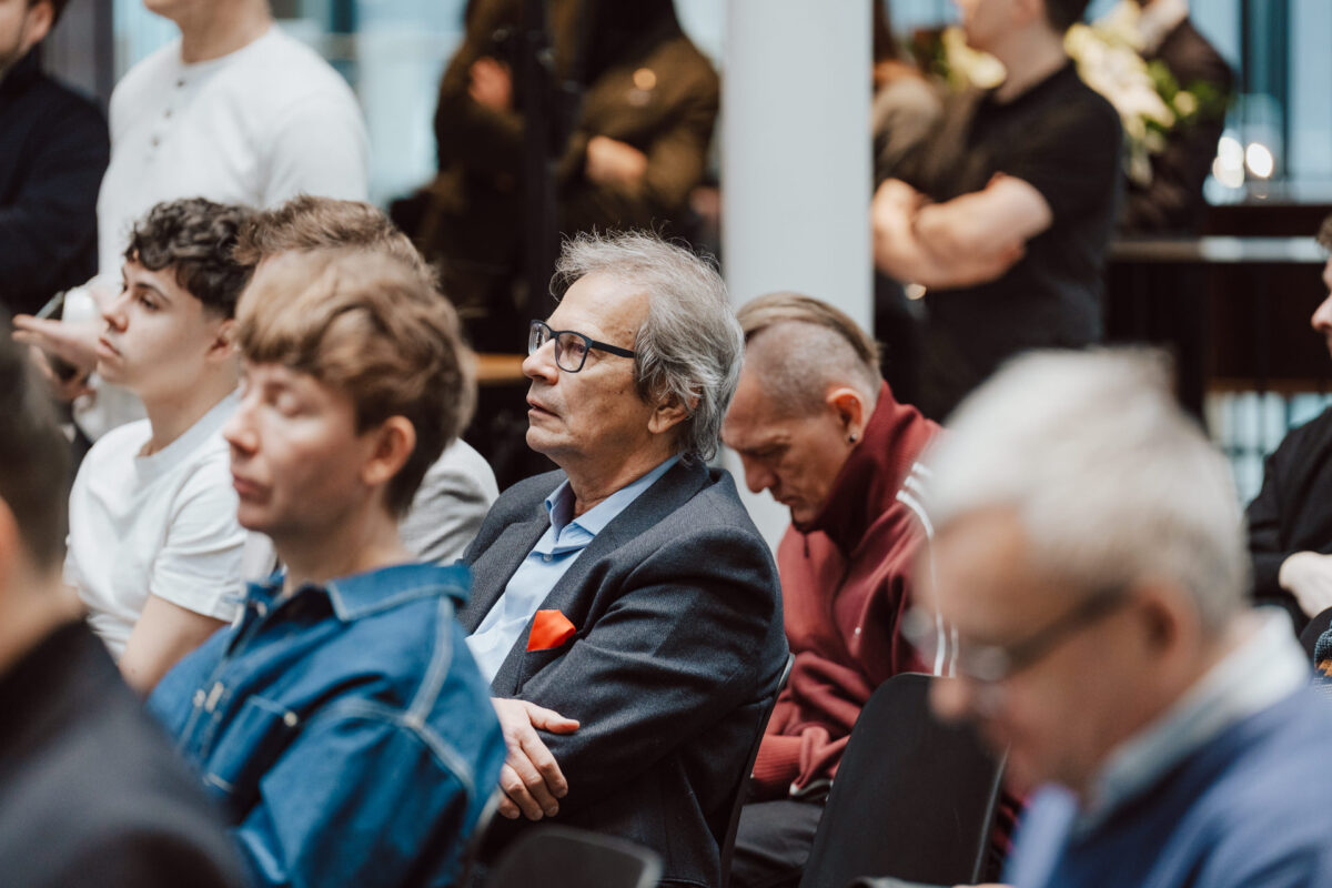 Eine Gruppe von Menschen, die in einem Raum sitzen und jemandem außerhalb des Rahmens aufmerksam zuhören. Die meisten scheinen konzentriert zu sein; eine Person in einer roten Jacke schaut nach unten. Der professionelle Rahmen lässt vermuten, dass es sich um eine Saloneröffnung oder ein Geschäftsseminar handeln könnte.  