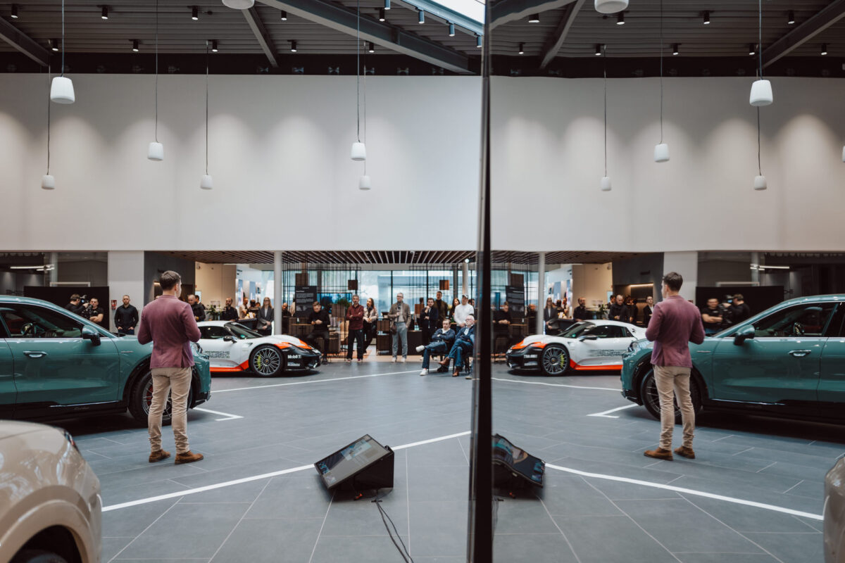 Ein Mann in einem rosafarbenen Hemd steht in einem modernen Autohaus während der Eröffnung des Ausstellungsraums und blickt dem Publikum zu. Sportwagen glänzen um ihn herum und ein zentraler Spiegel reflektiert die Szene und verdoppelt sein Bild, während sich im Hintergrund Menschen versammeln. 