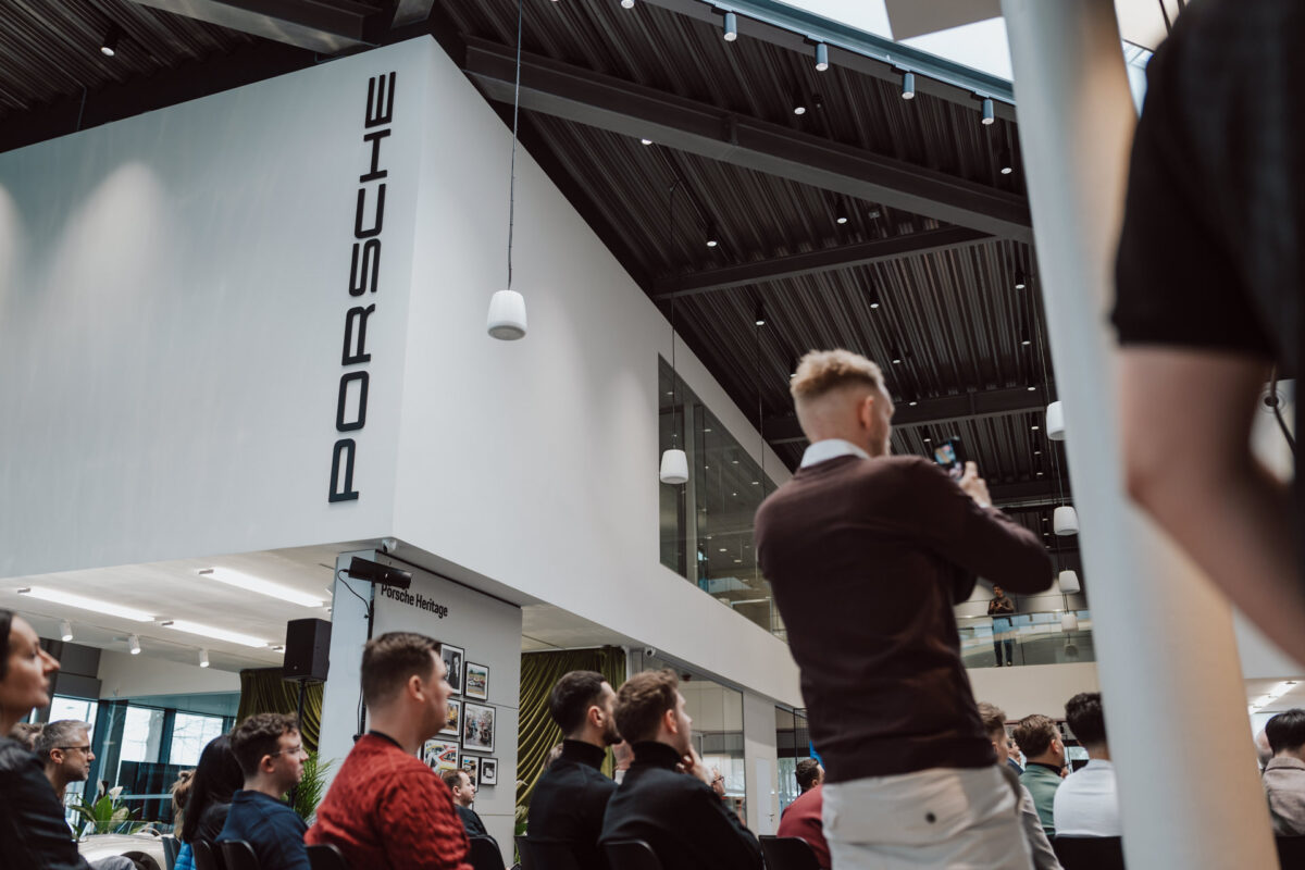 Die Menschen sitzen während der Eröffnung des Showrooms im modernen Porsche Showroom und hören aufmerksam zu, während eine Person steht, um ein Foto zu machen. Die hohen Decken des Showrooms und viel natürliches Licht schaffen eine einladende Atmosphäre für die Veranstaltung. 