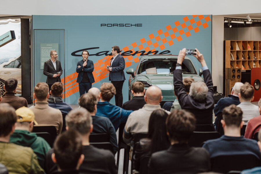 Trzech mężczyzn w garniturach stoi na scenie przed wystawą Porsche Cayenne w otwartym salonie, przemawiając do siedzącej publiczności; jeden z widzów robi zdjęcie smartfonem.
