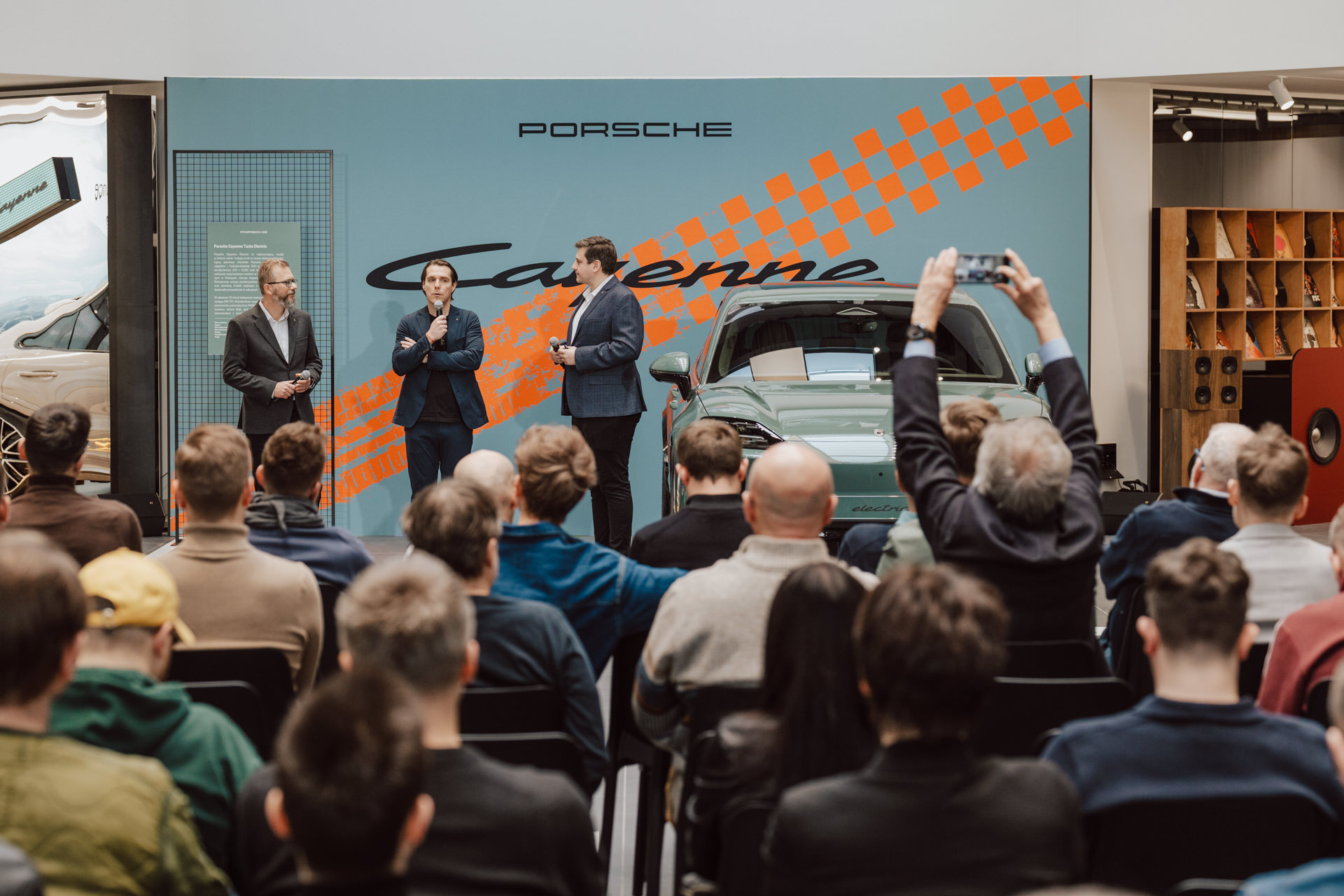 Trzech mężczyzn w garniturach stoi na scenie przed wystawą Porsche Cayenne w otwartym salonie, przemawiając do siedzącej publiczności; jeden z widzów robi zdjęcie smartfonem.