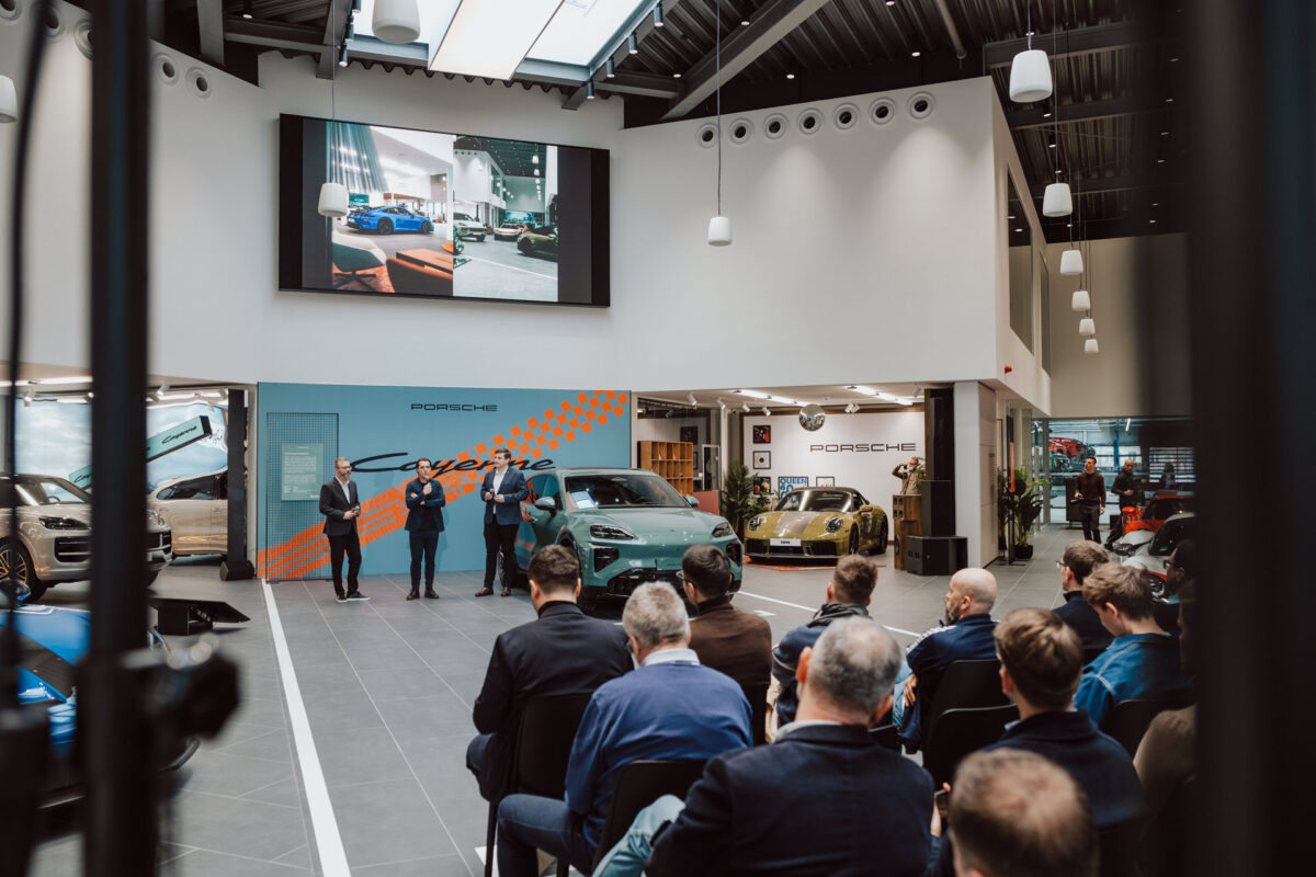 Eine Gruppe von Menschen sitzt vor einer Bühne in einem modernen Ausstellungsraum während der Eröffnung des Ausstellungsraums, in dem mehrere Porsche-Fahrzeuge ausgestellt sind. Vier Redner stehen an der Front, während Bilder der Autos auf eine große Leinwand über ihnen projiziert werden. 