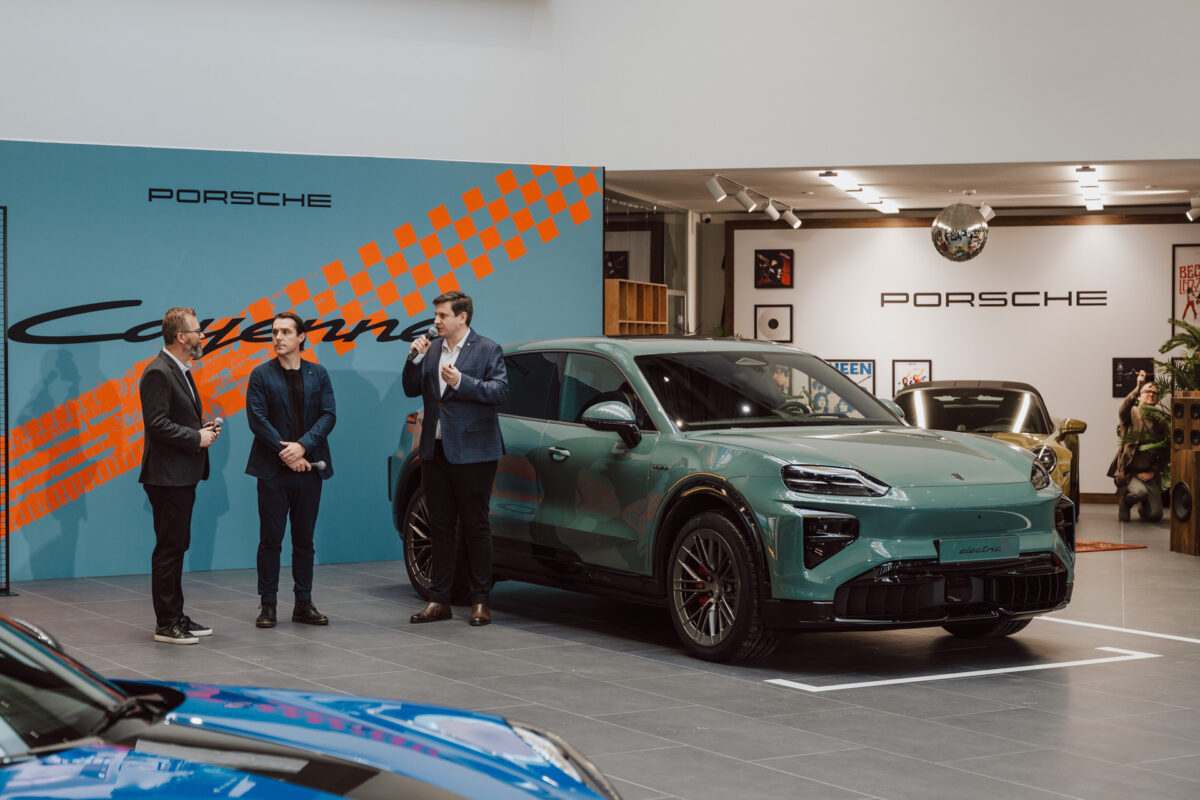 Drei Männer in Anzügen stehen neben einem grünen Porsche-Geländewagen in einem modernen Ausstellungsraum. An der Wand hinter ihnen prangt das Porsche-Branding und ein orangefarbenes Schachbrett, das die Eröffnung des Ausstellungsraums markiert.