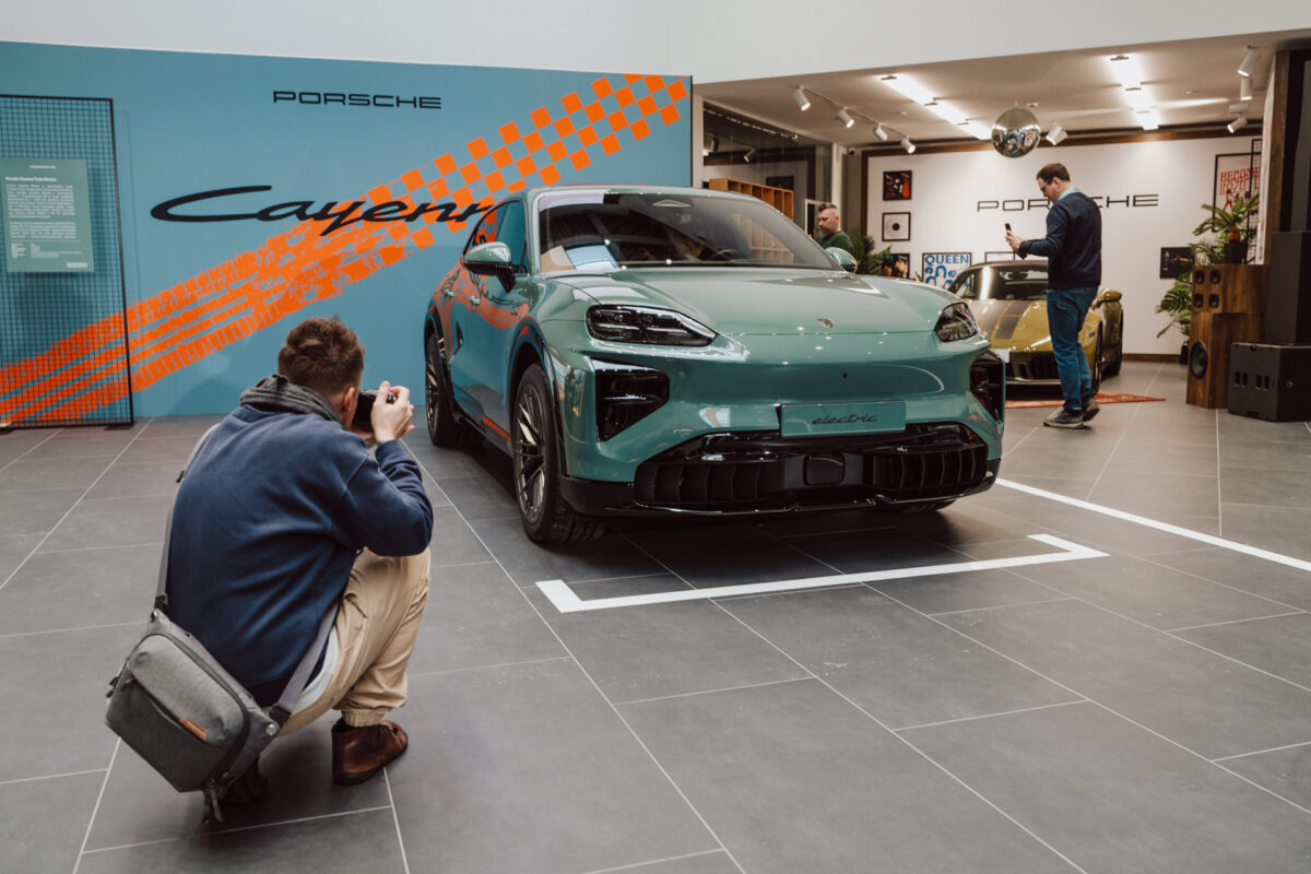 Eine Person geht in die Hocke, um einen türkisfarbenen Porsche zu fotografieren, der während der Eröffnung eines Showrooms in einem Raum ausgestellt ist. Eine andere Person steht im Hintergrund neben einem goldenen Porsche. Die Umgebung ist mit Porsche Markenzeichen und modernem Dekor ausgestattet. 