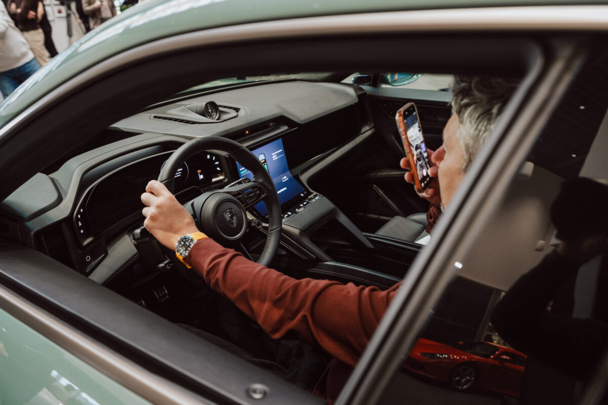 Eine Person sitzt auf dem Fahrersitz des Wagens, hält mit einer Hand das Lenkrad und nimmt mit einem Smartphone ein Foto oder ein Video auf - vielleicht um die Höhepunkte der Eröffnung des Ausstellungsraums festzuhalten. Das moderne Armaturenbrett und die Bedienelemente des Autos sind zu sehen. 