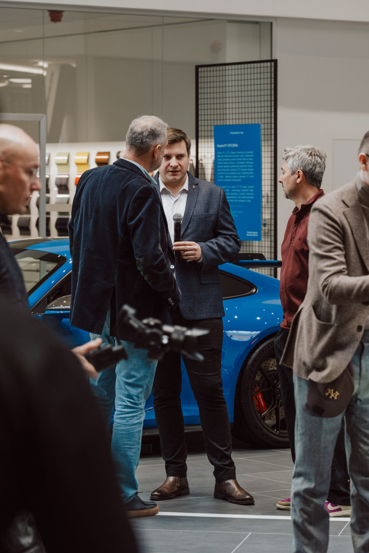 Während der Eröffnung des Showrooms spricht ein Mann im Anzug, der ein Mikrofon in der Hand hält, mit einem anderen Mann vor einem blauen Sportwagen im Showroom, während mehrere andere Personen in der Nähe sprechen.