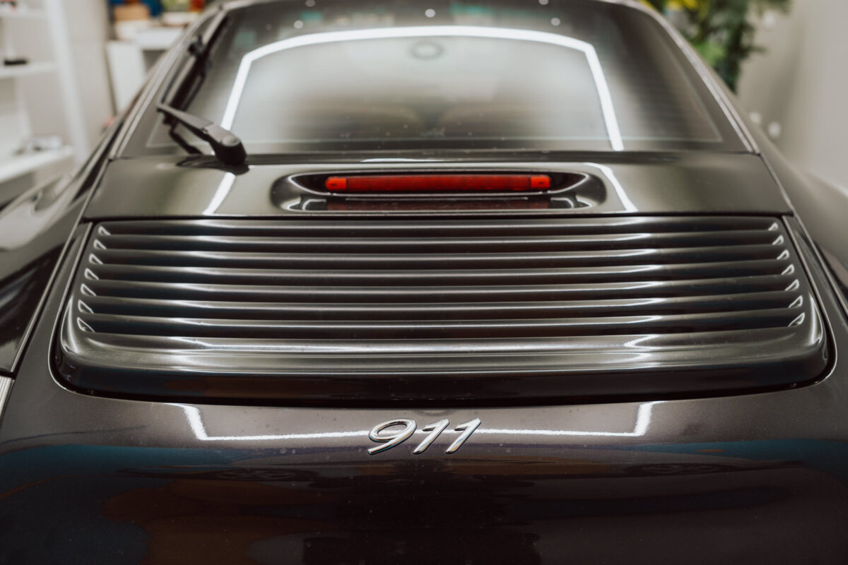 Nahaufnahme des Hecks eines dunklen Porsche 911 bei der Eröffnung des Showrooms. Zu sehen sind der hintere Kühlergrill, die Bremsleuchte und das '911'-Emblem, mit Reflexionen der Innenbeleuchtung auf der Oberfläche des Autos.