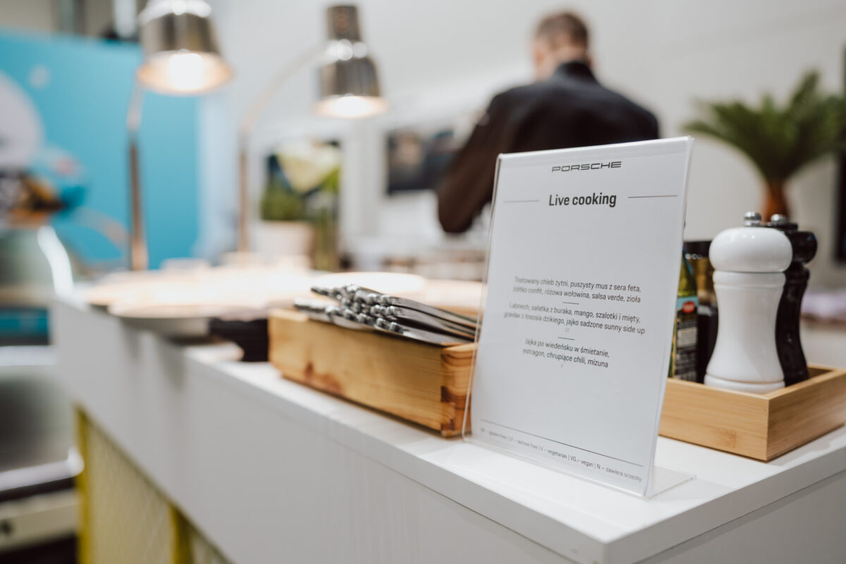 Der Schriftzug 'Live Cooking' steht auf einer weißen Arbeitsplatte mit Besteck, Salz- und Pfefferstreuern und Tabletts - und fängt die Spannung ein, die beim Öffnen des Wohnzimmers entsteht, wenn eine Person vor dem unscharfen, modernen Hintergrund der Küche arbeitet.