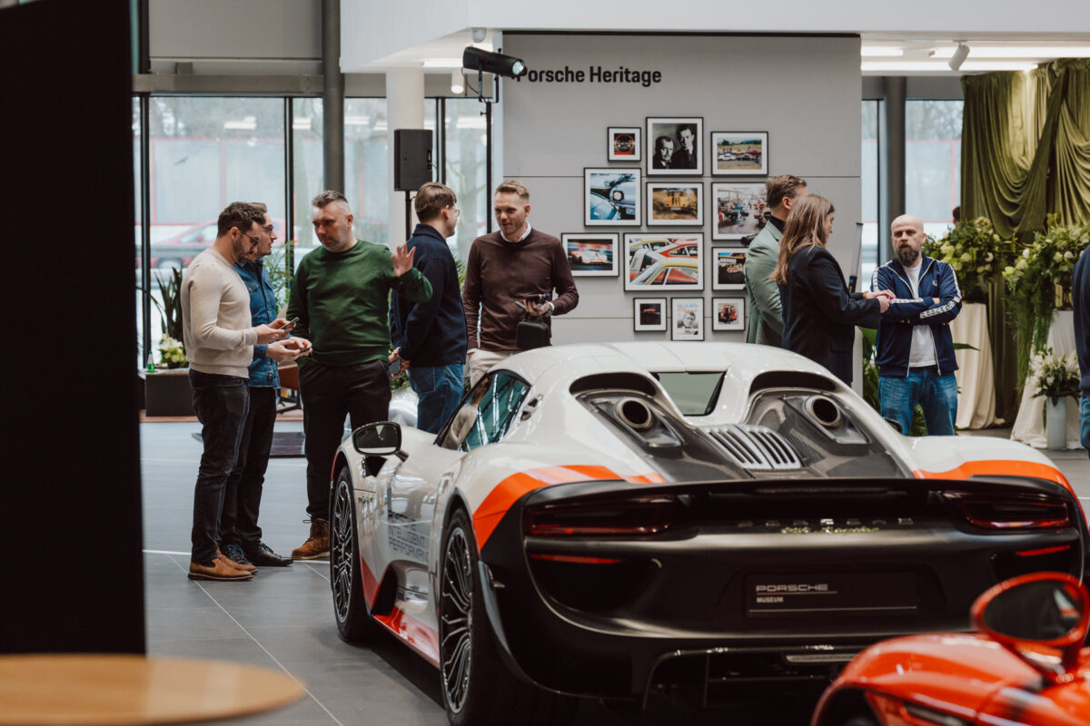 Eine Gruppe von Menschen plaudert und versammelt sich um einen schnittigen Porsche-Sportwagen während der Eröffnung eines Showrooms im Ausstellungsraum, mit Automobilfotos an der Wand und der Aufschrift 'Porsche Heritage' im Hintergrund.