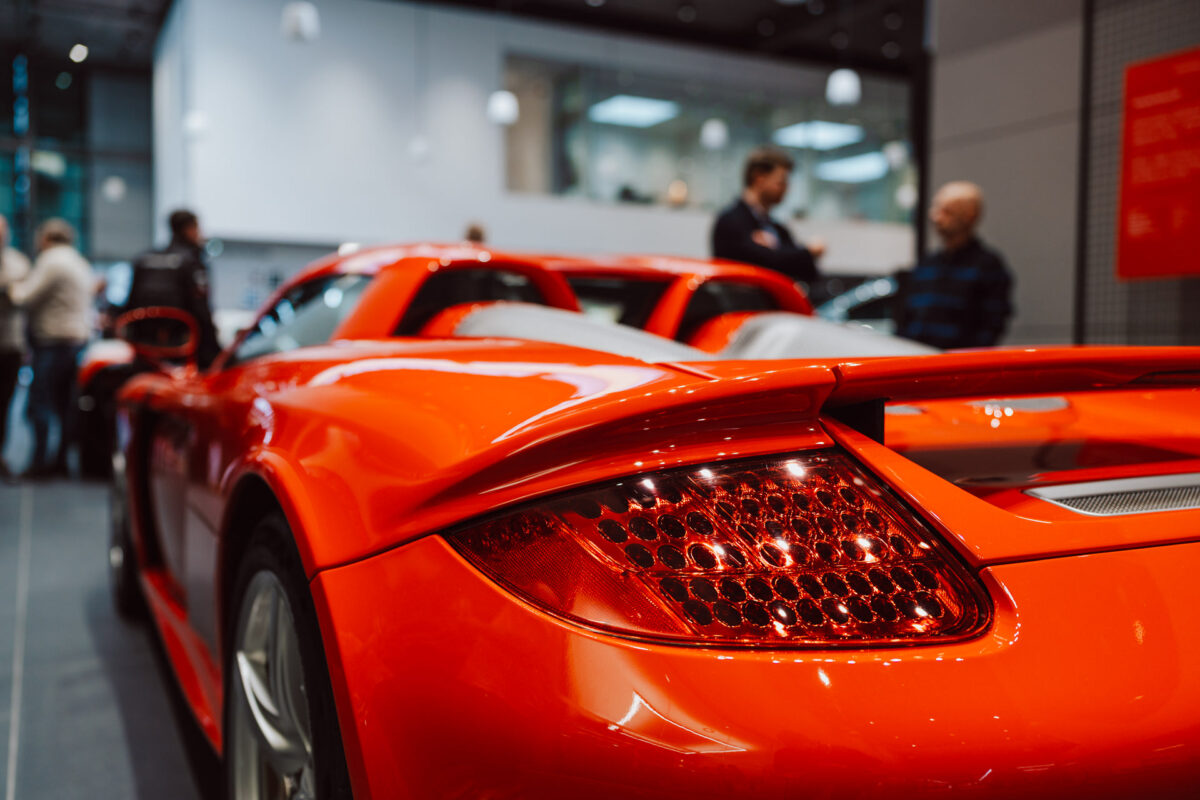 Nahaufnahme der Rückseite eines leuchtend roten Sportwagens, der in einem offenen Ausstellungsraum ausgestellt ist, mit Menschen im unscharfen Hintergrund und moderner Beleuchtung darüber, die eine lebhafte Ausstellungsraumatmosphäre schafft.