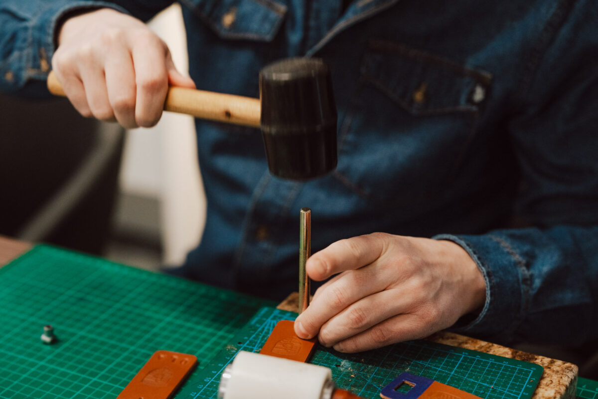 Die Person im Jeanshemd bearbeitet das Leder mit einem schwarzen Hammer und einem Metallwerkzeug auf einer grünen Schneidematte und fertigt das Design sorgfältig an - die perfekte Inspiration für einen Eröffnungssalon, in dem handgefertigte Details zählen.